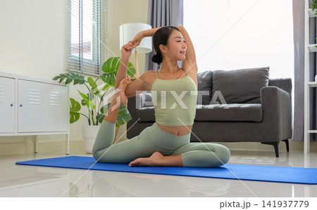 Sporty woman in green top and leggings stretching in Eka Pada Rajakapotasana exercise, One Legged King Pigeon pose on blue mat in bedroom at home. Sporty woman in green top and leggings stretching in Eka Pada Rajakapotasana exercise, One Legged King Pigeon pose on blue mat in bedroom at home. 141937779