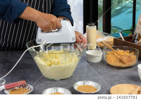 Baker using electric hand mixer to whip butter and cream in glass bowl and preparing homemade cake or dessert crust on dark kitchen counter Baker using electric hand mixer to whip butter and cream in glass bowl and preparing homemade cake or dessert crust on dark kitchen counter 141937781