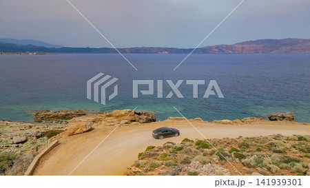Black car driving on dirt coastal road along sea with rocky shore and mountains in distance road trip travel exploration concept Greece landscape photo Black car driving on dirt coastal road along sea with rocky shore and mountains in distance road trip travel exploration concept Greece landscape photo 141939301