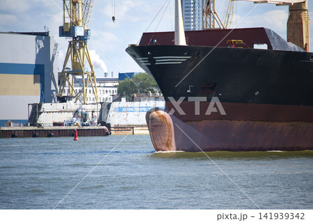 Cargo Ship Bow Passing a Busy Industrial Port With Cranes and Shipyard Infrastructure Cargo Ship Bow Passing a Busy Industrial Port With Cranes and Shipyard Infrastructure 141939342