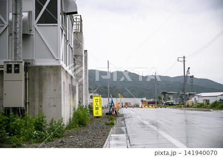 曇り空の下で雨に濡れた路面と立入禁止の看板の奥に建設途中の防潮堤が続く風景 141940070