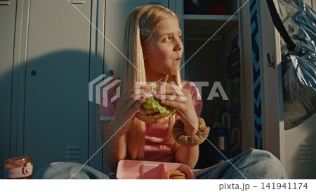 Young Schoolgirl Eating Homemade Sandwich from Reusable Pink Food Container Near Personal Locker 141941174