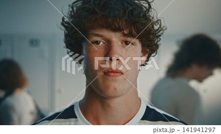 Dark Haired Curly Teenage Boy With Serious Facial Expression, Standing in Bright High School Hallway Dark Haired Curly Teenage Boy With Serious Facial Expression, Standing in Bright High School Hallway 141941297