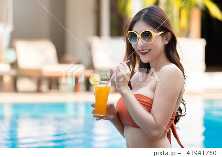 Sexy young asian woman in swimsuits in summer. beautiful woman in orange bikini with hat and sunglasses sitting in swimming pool with tropical drink. Sexy young asian woman in swimsuits in summer. beautiful woman in orange bikini with hat and sunglasses sitting in swimming pool with tropical drink. 141941780