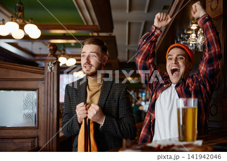 Pair of men demonstrate distinct emotional expressions while watching sports game on television Pair of men demonstrate distinct emotional expressions while watching sports game on television 141942046