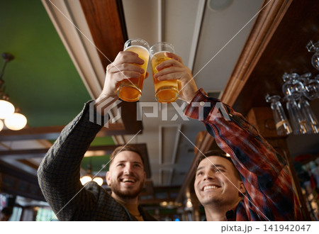 Two cheerful caucasian males celebrate with raised beer glasses and bright pub reflections Two cheerful caucasian males celebrate with raised beer glasses and bright pub reflections 141942047