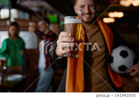 Man extending pint glass toward camera, soccer ball tucked under arm Man extending pint glass toward camera, soccer ball tucked under arm 141942058