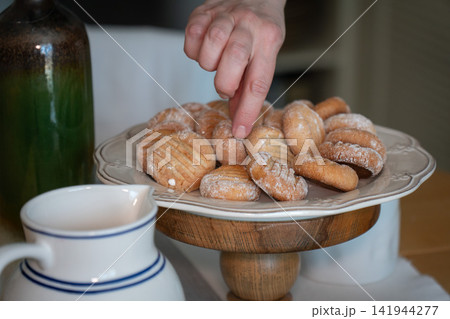 Female hand reaches for homemade almond cookie dusted with powdered sugar from cake stand. Home baking, dessert, snacking, hospitality, food styling, comfort, family tradition, lifestyle Female hand reaches for homemade almond cookie dusted with powdered sugar from cake stand. Home baking, dessert, snacking, hospitality, food styling, comfort, family tradition, lifestyle 141944277