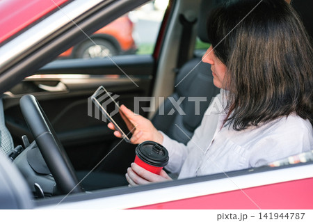 Woman driver sitting in car, holding a smartphone and red coffee cup, checking messages or navigating during a commute. Concept of modern urban lifestyle, multitasking, daily routine Woman driver sitting in car, holding a smartphone and red coffee cup, checking messages or navigating during a commute. Concept of modern urban lifestyle, multitasking, daily routine 141944787