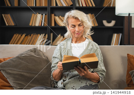 Comfort relaxed senior old elderly woman reading book at home. Mature woman resting enjoy calm day sitting on sofa at home alone. Adult lady spending free time keeping book smiling. Pension concept Comfort relaxed senior old elderly woman reading book at home. Mature woman resting enjoy calm day sitting on sofa at home alone. Adult lady spending free time keeping book smiling. Pension concept 141944918