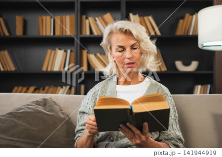 Comfort relaxed senior old elderly woman reading book at home. Mature woman resting enjoy calm day sitting on sofa at home alone. Adult lady spending free time keeping book smiling. Pension concept Comfort relaxed senior old elderly woman reading book at home. Mature woman resting enjoy calm day sitting on sofa at home alone. Adult lady spending free time keeping book smiling. Pension concept 141944919