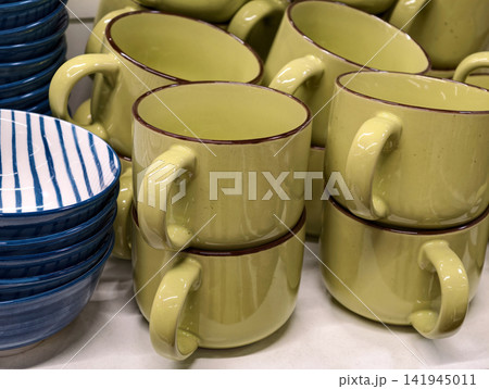 Ceramic cups in yellow green tones combined with stacked blue deep plates displayed in supermarket shelving forming simple kitchenware collection for daily household use and retail sale Ceramic cups in yellow green tones combined with stacked blue deep plates displayed in supermarket shelving forming simple kitchenware collection for daily household use and retail sale 141945011