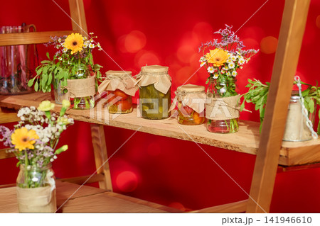 Jars filled with pickles and flowers are placed on a wooden shelf against a bright red background at a market event Jars filled with pickles and flowers are placed on a wooden shelf against a bright red background at a market event 141946610