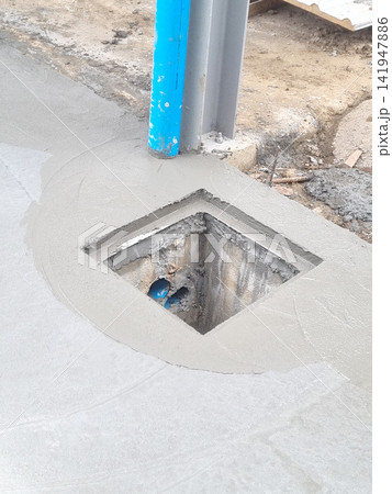 Freshly poured wet concrete floor at a construction site featuring a square drain opening, a blue utility pipe, and a grey structural steel beam. Industrial infrastructure development process. Freshly poured wet concrete floor at a construction site featuring a square drain opening, a blue utility pipe, and a grey structural steel beam. Industrial infrastructure development process. 141947886