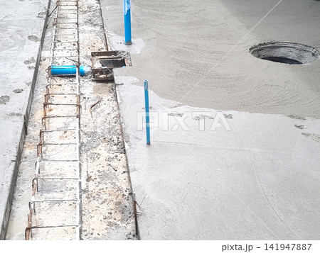 Wet concrete being poured at a construction site with steel rebar reinforcement, blue PVC drainage pipes, and manhole access. Professional industrial floor installation and civil engineering work. Wet concrete being poured at a construction site with steel rebar reinforcement, blue PVC drainage pipes, and manhole access. Professional industrial floor installation and civil engineering work. 141947887