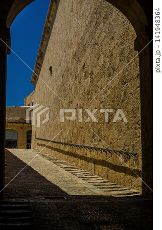 Dramatic archway frames a sunlit limestone staircase along Malta's ancient fortress walls, contrasting deep shadows with golden stone. . High quality photo 141948364