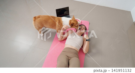 Cheerful Asian woman lying on exercise mat laughing while Corgi dog licks her face at home 141951132