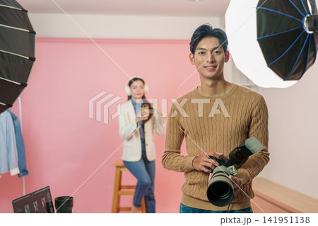 Asian male photographer holding camera in studio during photoshoot with model Asian male photographer holding camera in studio during photoshoot with model 141951138
