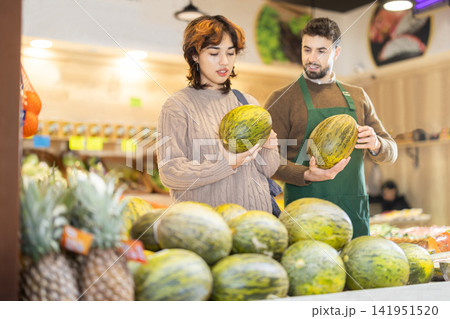 Girl customer in vegetable store examines melon, male seller advises and help with choice Girl customer in vegetable store examines melon, male seller advises and help with choice 141951520