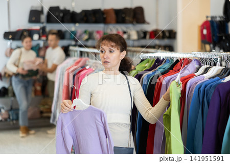 Woman buyer in store has removed hanger with blouse from window and is considering product. Woman buyer in store has removed hanger with blouse from window and is considering product. 141951591