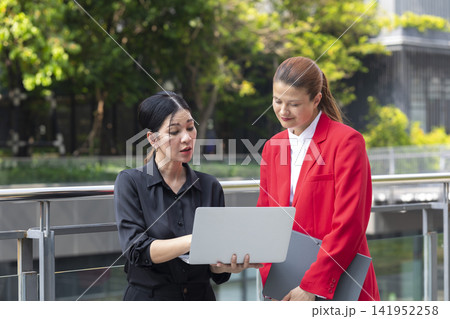 Asian woman and Caucasian woman in suit talking and working with laptop computer in outdoor office space. Successful and teamwork concept. 141952258