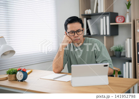 Thoughtful man wearing glasses sitting at wooden desk looking at tablet screen with hand on face in bright room with bookshelf and natural light 141952798