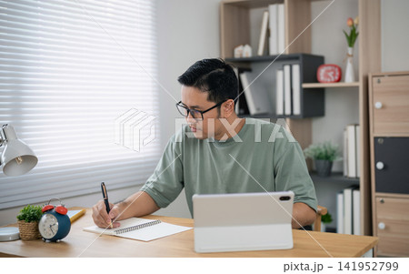 Young man wearing glasses writing notes with pen while using tablet computer at home office desk surrounded by modern decor and natural light window 141952799