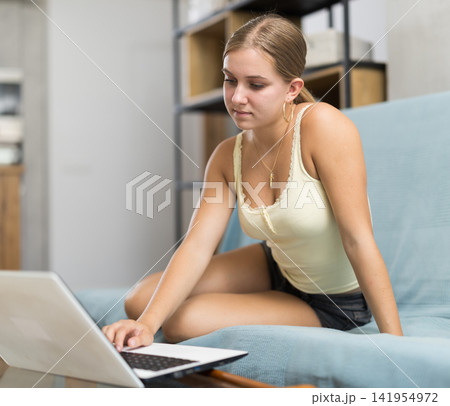 Teenage girl working on laptop while sitting on cozy sofa Teenage girl working on laptop while sitting on cozy sofa 141954972