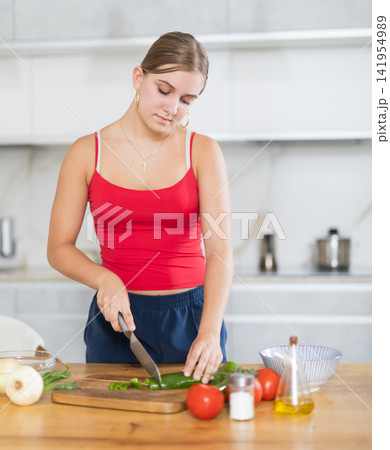 Teenage girl standing in kitchen and preparing salad Teenage girl standing in kitchen and preparing salad 141954989