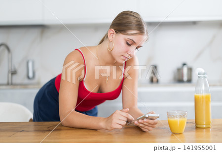 Teenage girl looking at smartphone while drinking fresh juice in kitchen 141955001