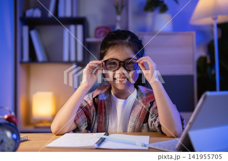 Happy young girl sitting at desk adjusting large glasses while doing homework with open book and digital tablet in cozy evening room Happy young girl sitting at desk adjusting large glasses while doing homework with open book and digital tablet in cozy evening room 141955705