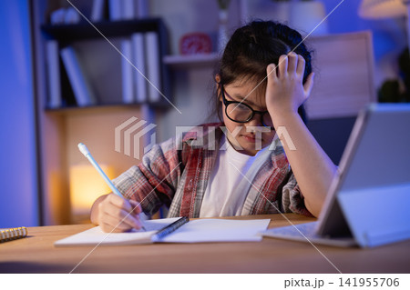 Young girl with glasses struggling to concentrate on homework studying late at wooden desk with tablet and notebook at home in cozy room with warm lighting Young girl with glasses struggling to concentrate on homework studying late at wooden desk with tablet and notebook at home in cozy room with warm lighting 141955706