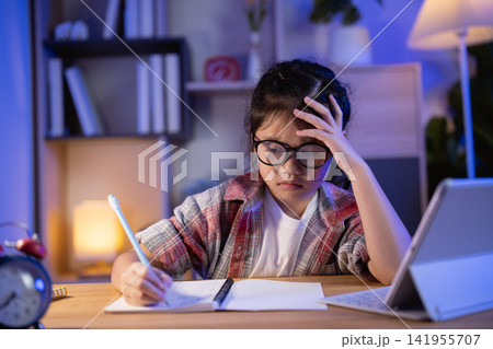 Young girl with glasses thinking hard while studying late at night with laptop and notebook in cozy room lighting and focused expression Young girl with glasses thinking hard while studying late at night with laptop and notebook in cozy room lighting and focused expression 141955707
