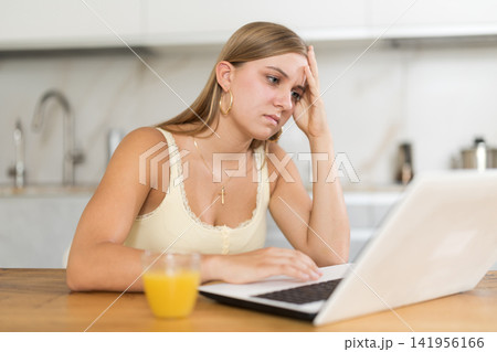 teenager is sitting at a table in the kitchen and sadly looking at a laptop screen teenager is sitting at a table in the kitchen and sadly looking at a laptop screen 141956166