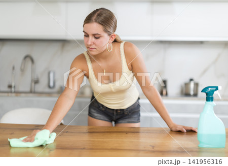 Teenage girl wiping wooden surfaces with cleaner in kitchen Teenage girl wiping wooden surfaces with cleaner in kitchen 141956316