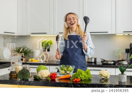 Young woman dancing with headphones while mixing fresh salad for healthy keto diet in home kitchen Young woman dancing with headphones while mixing fresh salad for healthy keto diet in home kitchen 141956395