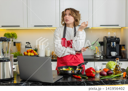 Young woman cooking fresh vegetables while looking at laptop in home kitchen for healthy diet meals 141956402
