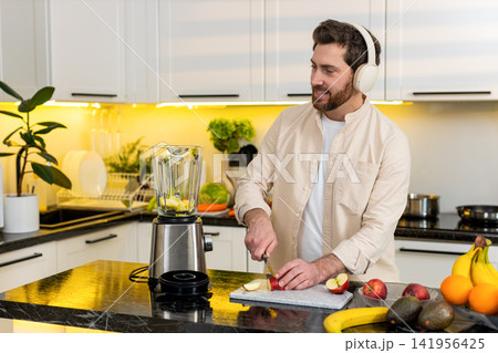 Middle-aged man making apple smoothie in kitchen listening to music on headphones for healthy diet Middle-aged man making apple smoothie in kitchen listening to music on headphones for healthy diet 141956425