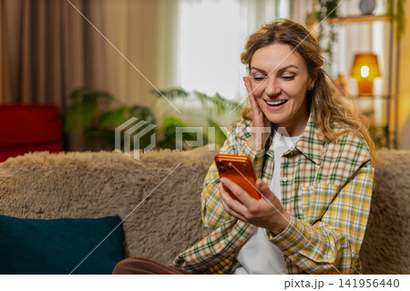 Young woman on home sofa reading delivery message on smartphone celebrating online game win with joy Young woman on home sofa reading delivery message on smartphone celebrating online game win with joy 141956440