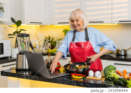 Senior old woman cooking vegetables in kitchen using laptop recipe for healthy diet home lifestyle 141956444
