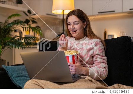 Adult woman with laptop and popcorn watches trailer movie online laughs comments comedy fun at night Adult woman with laptop and popcorn watches trailer movie online laughs comments comedy fun at night 141956455