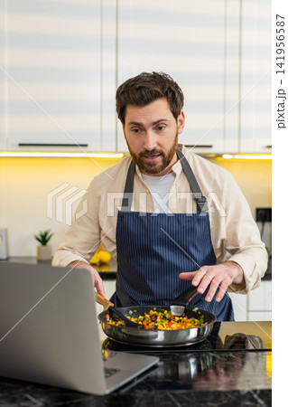 Middle-aged man cooking vegetables on skillet while live streaming diet recipe on laptop in kitchen Middle-aged man cooking vegetables on skillet while live streaming diet recipe on laptop in kitchen 141956587