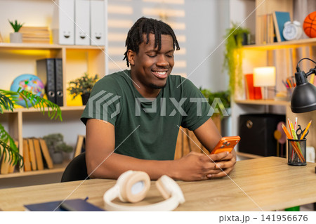 African American young man on home table smiling at smartphone message during relaxed online chats African American young man on home table smiling at smartphone message during relaxed online chats 141956676