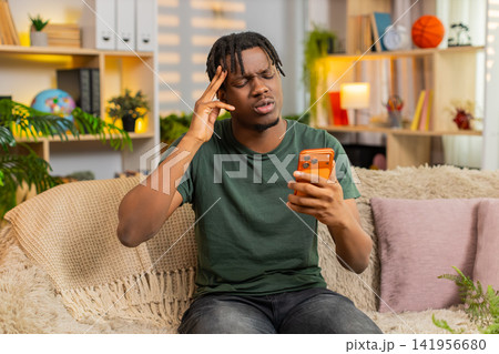 African American young man on home sofa feeling upset at bad smartphone news after online game loss African American young man on home sofa feeling upset at bad smartphone news after online game loss 141956680