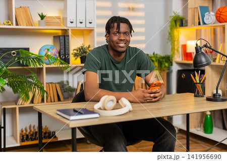 African American young man on home table smiling at smartphone message during relaxed online chats African American young man on home table smiling at smartphone message during relaxed online chats 141956690