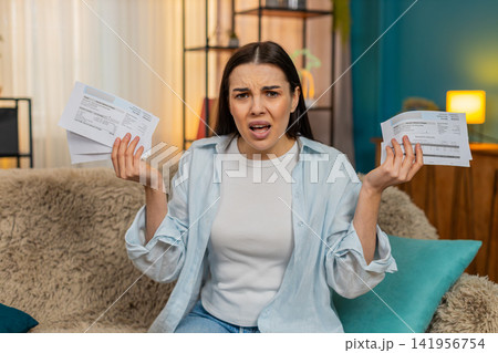 Young woman disappointed stressed sits on sofa holding bills worried financial problems asking why 141956754