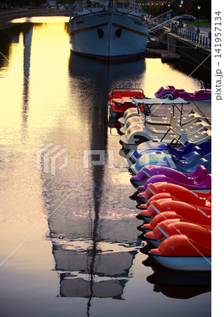 Colorful Pedal Boats Docked Along Canal With Large Ship and Golden Sunset Reflections Colorful Pedal Boats Docked Along Canal With Large Ship and Golden Sunset Reflections 141957134
