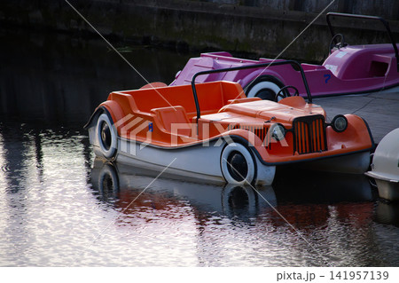 Vintage Orange Pedal Boat Docked on Calm Water With Retro Car Design and Reflections Vintage Orange Pedal Boat Docked on Calm Water With Retro Car Design and Reflections 141957139
