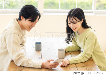 スマートフォンを一緒に操作し画面を見ながら笑顔で会話する30代夫婦 スマートフォンを一緒に操作し画面を見ながら笑顔で会話する30代夫婦 141962437