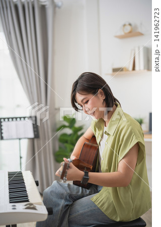 Woman playing acoustic guitar in bright modern living room with keyboard and bookshelf, relaxing and enjoying music 141962723